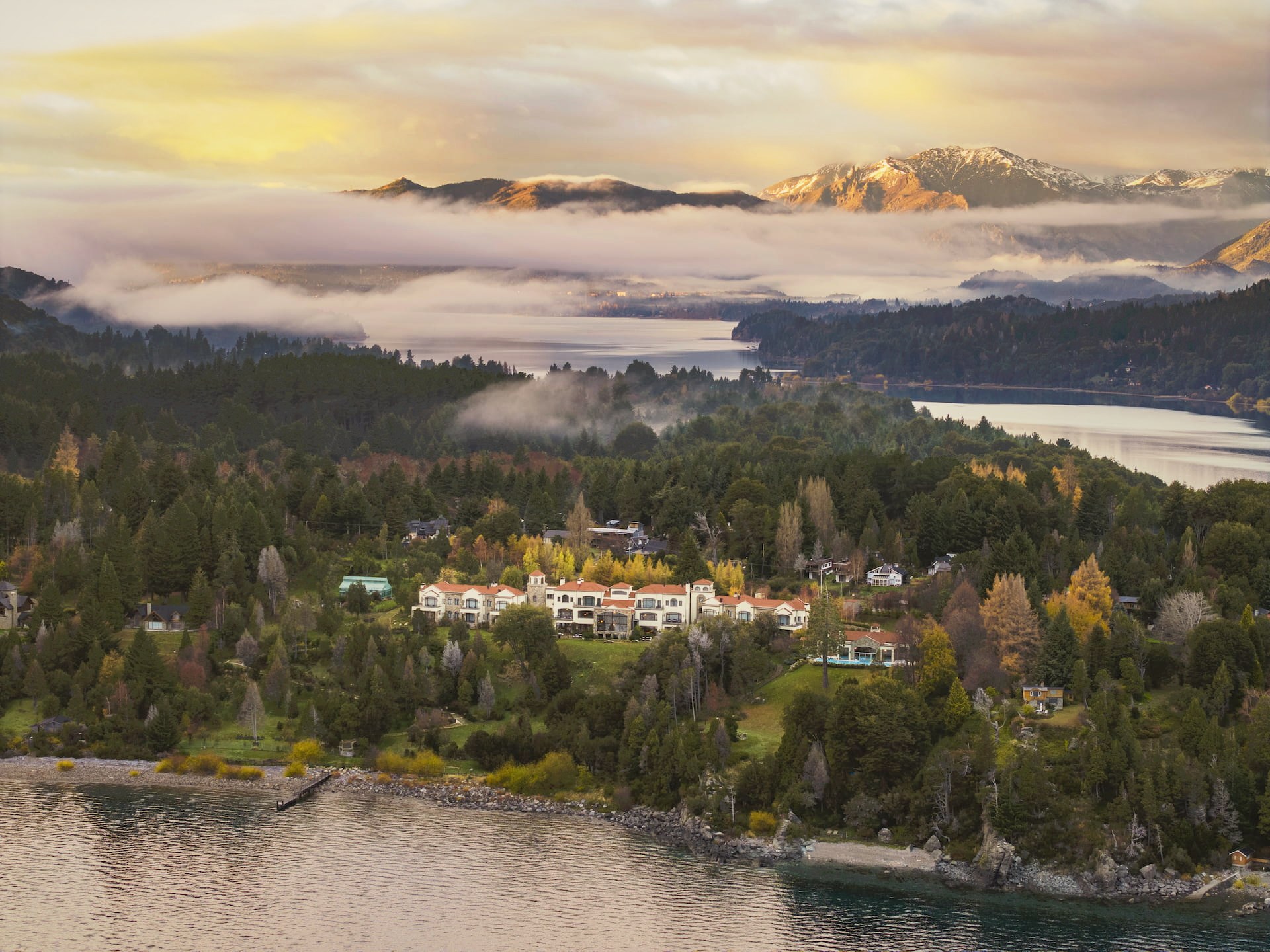 Aerial sunrise view of Villa Beluno surrounded by autumn Patagonian forest on the shores of Nahuel Huapi Lake with snow-capped Andes in the background, Bariloche, Argentina