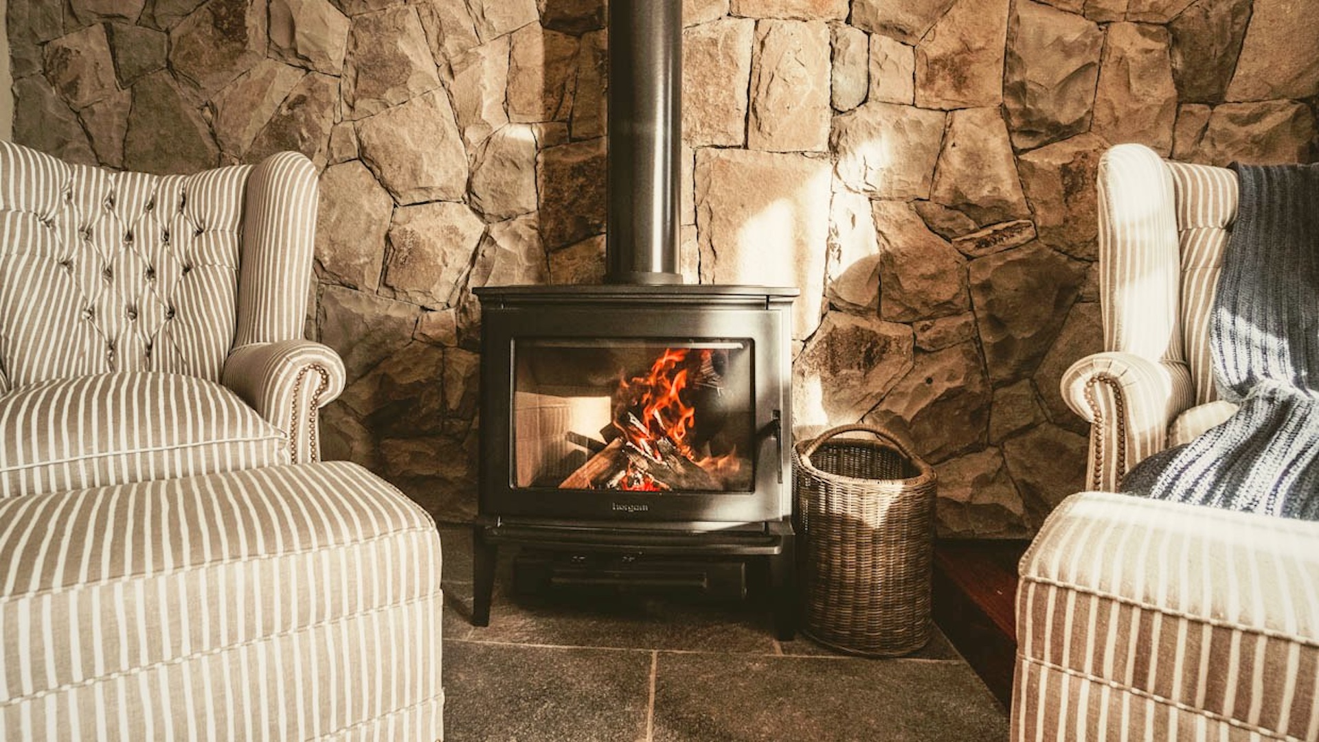 Dos sillones de tela a rayas frente a una chimenea de leña encendida junto a pared de piedra patagónica en Villa Beluno, hotel boutique de lujo en Bariloche, Patagonia