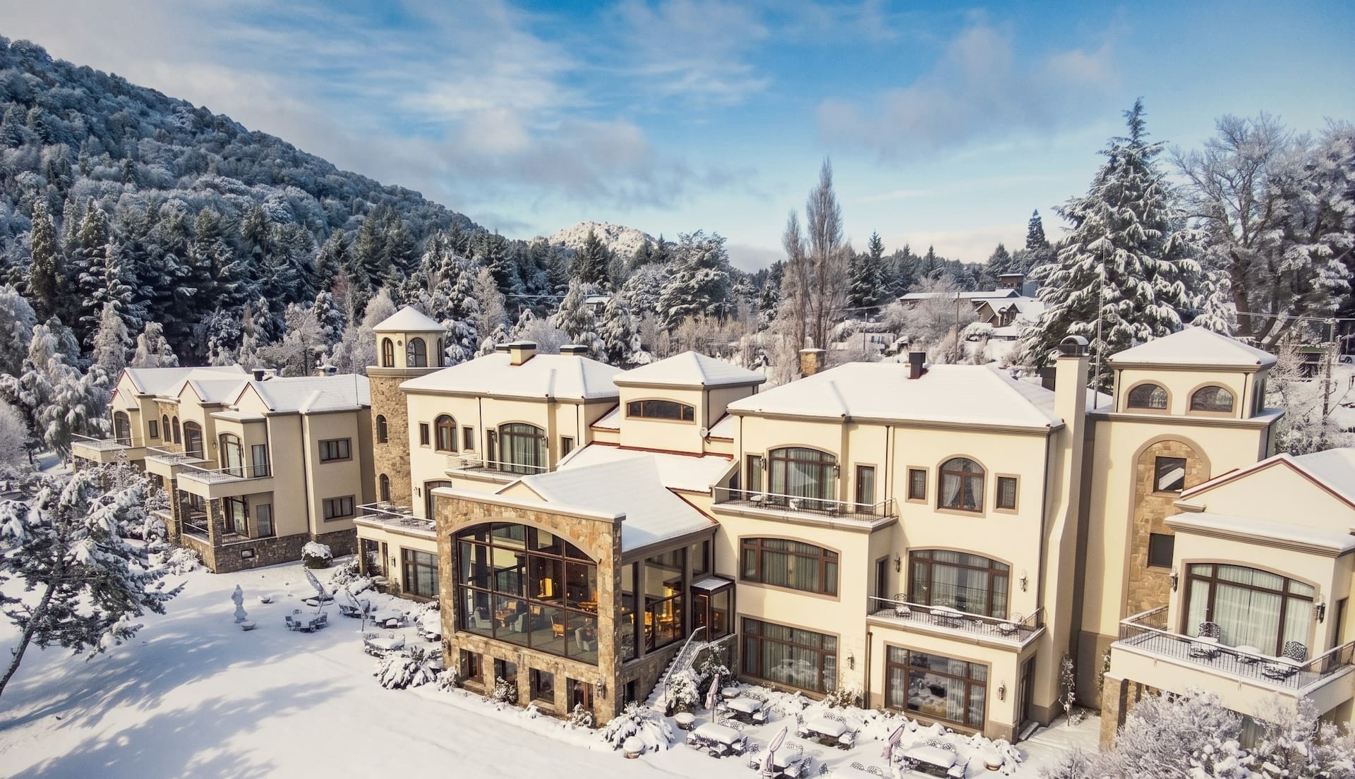 Vista aérea de Villa Beluno cubierto de nieve en invierno con el gran salón acristalado iluminado y el bosque patagónico nevado alrededor, Bariloche, Argentina
