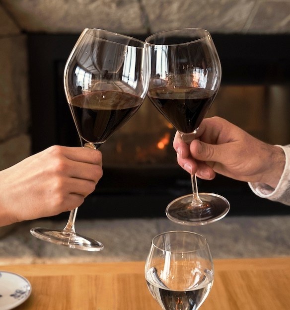 Dos manos brindando con copas de vino tinto frente a una chimenea encendida en Villa Beluno, hotel boutique en Bariloche, Río Negro, Argentina